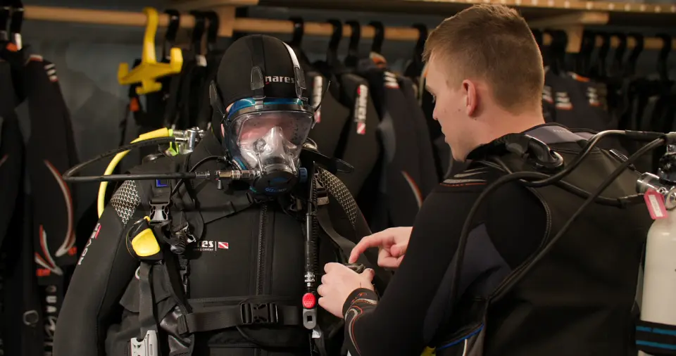 A diver being fitted with a full-face mask and regulator in the kit room