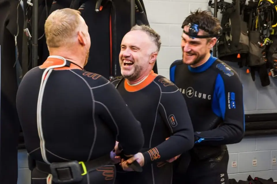 Three people in wetsuits laughing together in the changing room before a dive